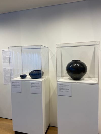 The image shows two ceramic pieces on display at the Bruce Museum in Greenwich, Connecticut. The pieces are displayed in two separate, clear acrylic cases, each resting on a white pedestal. The case on the left contains two pieces. The smaller piece is a black bowl and the larger piece is a black vase. The case on the right contains a single piece, a larger black vase. All three pieces appear to be made of black pottery, and each has an inscription next to it providing more details about the pieces. The museum walls are white, and the floor appears to be made of wood. There are no people in the scene.