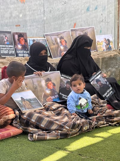This image, captured in Dura, Palestinian Territory, depicts four individuals seated outdoors on artificial green turf, with a light-colored concrete wall in the background. A brown and beige plaid blanket covers a significant portion of the foreground. On the left, a young boy in a light-colored t-shirt holds a portrait of a young man in a yellow hoodie. In the center foreground, an infant wearing a light blue long-sleeved shirt with a panda graphic sits upright, looking directly at the camera. Behind them, two adult women, both wearing black niqabs that obscure their faces except for their eyes, are seated. The woman on the left holds a large portrait of a young man in a red cap and glasses, holding a lit sparkler. The woman on the right holds a different portrait of a young man, featuring the text "JUSTICE FOR AWDEH" in English and "צדק לעודה" (Justice for Awdeh) in Hebrew. Several additional copies of the portrait showing the young man with the sparkler are propped against the concrete wall in the background. Some of these posters bear text in English ("JUSTICE FOR AWDAH"), Arabic ("احترام الشهيد" - Respect the martyr), and Hebrew ("כבוד המת" - Honor the dead, and "צדק לעודה"). A smaller portrait of an adult holding a child is also visible against the wall. The wall itself has faint markings and Arabic script, including the word "Dura". Two patterned pillows are visible on the far left, partially covered by the blanket. Natural daylight illuminates the scene.