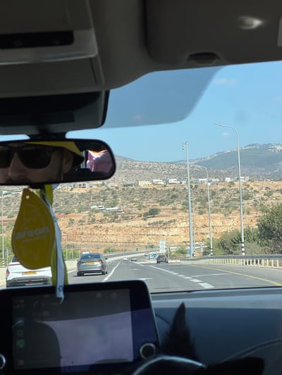 The image depicts a daytime scene from the interior of a vehicle traveling on a multi-lane highway within the Merom HaGalil Regional Council, Israel.
In the foreground, a portion of the vehicle's dashboard and infotainment screen is visible, displaying a navigation interface. Hanging from the rearview mirror is a yellow "Areeon" brand car air freshener. The rearview mirror itself reflects a person, presumably the driver, wearing dark sunglasses and a yellow hat.
The midground shows the highway extending forward, with white lane markings. Several vehicles are ahead, traveling in the same direction. A white SUV with an Israeli license plate (361:43-101) is in the left lane, and a silver sedan with an Israeli license plate (332:01-601) is further ahead. To the right of the highway, a metal guardrail separates the road from the shoulder, where tall streetlights with modern fixtures are installed. One pole features what appears to be a surveillance camera. Road signs, including a blue circular sign indicating a mandatory right turn or direction, are visible on the right side of the road.
The background features arid, rocky hillsides characterized by terraced sections and sparse green vegetation, consistent with the Galilee region. Scattered white residential buildings are visible on the lower and mid-slopes of these hills. The sky above is clear and blue, indicating bright, sunny weather.