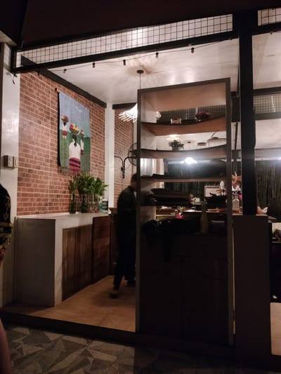The image captures an interior scene at night, likely a restaurant or cafe in Kidapawan City, Philippines. The foreground features patterned tile flooring transitioning to a wooden floor leading into the establishment. A person, whose lower body is visible, walks away from the camera into the darker interior.
The establishment has a rustic aesthetic, with a prominent brick wall adorned with a tapestry depicting three figures carrying baskets. Several green glass bottles containing plants are arranged on a counter. To the right, a tall, open shelf unit displays various items, including what appear to be books or folded materials, decorative bowls, and bottles. Behind the shelf unit, the environment becomes darker, with glimpses of bamboo-like structures and other patrons seated at tables. Overhead, simple string lights and a caged light fixture illuminate the space. The overall atmosphere is dimly lit and somewhat obscured by shadows.