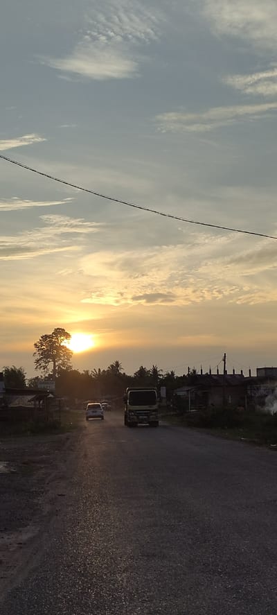 The image depicts a road scene in Blangmajron, Indonesia, during sunset. The sun is visible as a bright, luminous orb near the horizon, casting a warm, golden glow across the sky and illuminating the clouds with hues of orange and yellow. The sky is a gradient of pale blue at the top, transitioning to softer tones as it approaches the sunset. A power line stretches across the upper portion of the frame.
In the foreground, a paved road runs through the scene, with a white car and a truck driving away from the viewer. The road surface appears worn and textured. To the left of the road, there are some simple structures, possibly makeshift stalls or small buildings, with corrugated iron roofs. A tall, leafy tree stands prominently on the left side of the road, silhouetted against the bright sunset.
To the right of the road, more buildings are visible, some appearing to be under construction with exposed wooden frames and roofing materials. There is a hint of smoke or steam rising from a pile of debris on the far right. The landscape behind the road is dotted with trees, including palm trees, suggesting a tropical or sub-tropical environment. The overall impression is one of a rural or semi-urban setting in Indonesia as the day concludes.