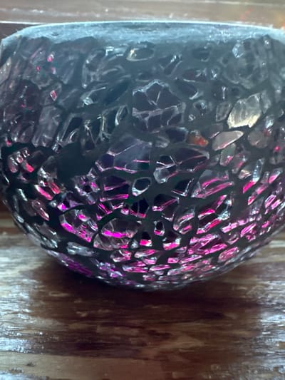 The image shows a close-up of a round, dark mosaic object with iridescent pink and clear glass fragments. The object appears to be a decorative vessel or lantern, possibly a candle holder. It is placed on a wooden surface, likely a table or shelf, which has a visible wood grain pattern. The lighting suggests the object is illuminated from within or by a nearby light source, causing the glass pieces to reflect and refract light in vibrant pink and shimmering clear tones. The background is out of focus, but appears to be a continuation of the wooden surface or a textured wall. There are no people or animals visible in the image, and no specific actions or interactions are depicted. The scene is static and focuses solely on the texture and reflective qualities of the mosaic object. No information can be extracted from this image regarding its location in Hradec-Nová Ves, Czechia.