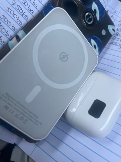 This is a close-up, overhead shot of a white magnetic wireless power bank resting on a blue-lined notebook. The power bank has a circular indentation on its surface with a lightning bolt logo in the center. To the right of the power bank, a white earbud charging case is visible. Partially in the background, a phone case with an evil eye design can be seen. The notebook pages appear to have handwritten notes, including numbers and "bs." The overall scene suggests a personal item setting, possibly a desk or table. The location in Conuco Viejo, Venezuela is not visually represented in this image.
