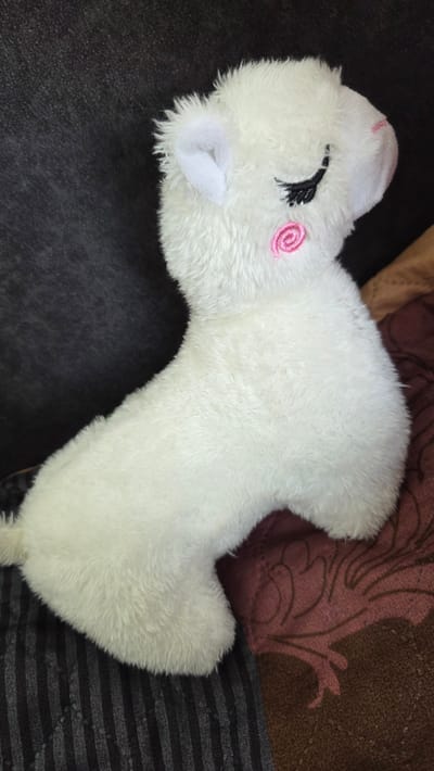 A white, plush unicorn toy rests on a surface. The toy features a closed eye with black stitching and a pink, swirled cheek marking. The surface beneath the toy appears to be a mix of textures, including dark gray and patterned brown fabrics.