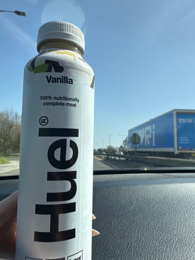 The image shows a bottle of Huel Vanilla flavor, described as "100% nutritionally complete meal," held in what appears to be a vehicle interior, likely a car based on the textured dashboard. Outside the window, a blue truck with "EVRI" branding is visible, along with highway structures like lampposts and possibly other vehicles. The sky is clear blue. It's difficult to confirm this location is specifically in Kettering, United Kingdom without further information or references visible in the photo beyond general road infrastructure.
