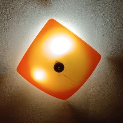 The image shows an interior scene featuring a ceiling light fixture. The fixture is square-shaped with rounded corners and has an orange-yellow translucent diffuser. Two bright light sources are visible within the diffuser, along with a dark, circular mounting point in the center. The ceiling itself appears textured and is illuminated unevenly by the light, with areas of bright light and shadows. The overall impression is of a ceiling light, possibly in a home or public space. There is no indication of the city of Lyubimets, Bulgaria, in the image.