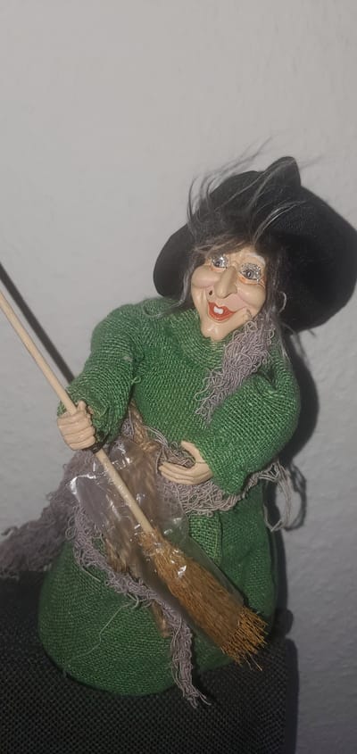 A doll of a witch is posed with a broom in front of a white wall. The witch wears a black hat, a green dress, and a gray scarf. She has a painted face with blue glitter on her eyelids, rosy cheeks, and red lips. Her hair is gray and windswept. The broom is made of straw and wood. The doll is positioned with its left arm bent and holding the broom. The background appears to be an indoor setting. There is no indication of the location being Sasbach, Germany.