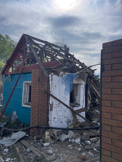 The image shows a destroyed building in Bilopillia, Ukraine. The building is a small, single-story structure with a blue wooden exterior and a white brick wall. The roof has been completely destroyed, and there are large pieces of debris scattered around the building. The building is located on a plot of land with green grass and a brick wall to the right. The debris includes wood, metal, and brick. The building is partially visible through a gap in the brick wall. The environment is overgrown with vegetation. There is a blue tarp or sheet of material in a window of the building. The sky is partially cloudy with a bright sun. The scene appears to be the aftermath of a bombing or other destructive event. The image was likely captured on a smartphone or other similar digital device. The image is grainy and the resolution is low.