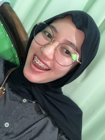An individual is depicted in an indoor setting, purportedly located in Patrang, Indonesia. The subject is a female with medium skin tone, wearing a dark grey patterned long-sleeved shirt and a black headscarf (hijab). She wears round, gold-rimmed glasses and has clear orthodontic braces visible on her upper teeth. She is smiling broadly, with her mouth open, and gazing directly at the camera. The background consists of a light green wall or fabric, with a portion of a dark brown object, possibly furniture with green upholstery and metal studs, visible on the left side.