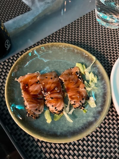 An overhead, close-up shot displays a meal served on a dark, woven placemat, indicative of a restaurant setting. The primary focus is a round, speckled blue-green ceramic plate featuring three portions of seared or marinated fish, possibly salmon or tuna. Each piece of fish is glazed with a dark, glossy sauce and liberally sprinkled with both black and white sesame seeds, resting atop a small bed of finely shredded light green lettuce.

The ceramic plate has a rustic appearance with a slightly raised, darker rim. In the upper left corner, a clear drinking glass is partially visible, reflecting light. A fragment of another lighter-colored dish or bowl can be seen in the upper right. The dark placemat underneath has a distinct, tightly woven or grid-like pattern in shades of dark grey and black. A blurred, dark object, potentially a bottle, is partially visible in the lower-left corner, though no clear text is discernible on it or elsewhere in the image. The scene suggests an indoor dining experience.