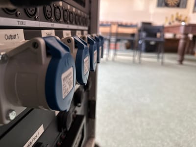 The image displays a close-up, angled view of rack-mounted electrical and audio equipment in the foreground, with a blurred background showing an indoor communal space. The equipment features several gray and blue industrial power sockets, manufactured by Mennekes, alongside multiple XLR audio connectors labeled "THRU" and "OUT," and text indicating "Output 1," "Output 2," and "Switch Pack 1." The background reveals a light-colored carpeted floor, numerous stacked and arranged wooden chairs, and a portion of a large wooden-legged object, possibly a pool table. A wall decoration, potentially a dartboard or tapestry, is faintly visible in the upper background. No individuals are present. The scene is located within a building in Whiteparish, United Kingdom.