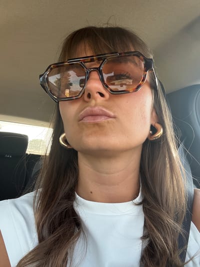 A medium close-up, rotated image, shows a person, likely female, with long, straight to slightly wavy brown hair, seated inside what appears to be a car. The image is oriented with the person's head on the left and their torso on the right, but describing it upright: the person is looking directly at the camera.

The subject is wearing a white t-shirt or top. Her skin has a tanned complexion. She wears oversized, square-shaped sunglasses with a tortoiseshell pattern, which are highly reflective. The reflections in the lenses show an outdoor scene with trees and possibly structures, indicating bright daylight. In the reflection of the left lens (viewer's right), visible eyelashes are prominent, hinting at her eye beneath the lens.

On her visible ear, she wears a thick, gold-colored ear cuff or hoop earring. Her lips are slightly parted, with a natural, neutral expression. Her long brown hair cascades over her shoulder and blends into the white fabric of her shirt.

The background suggests the interior of a car, with dark upholstery visible behind her head and possibly a seatbelt strap across her upper chest. Bright natural light streams in from what appears to be a car window, creating significant glare on the seat and around her hair. The time of day is clearly daylight, likely sunny given the strong reflections. There is no visible text in the image. The overall impression is a casual, relaxed moment captured in a car.