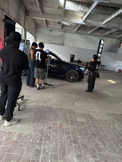 The scene appears to be a film or photography shoot taking place in a large, industrial-style building with exposed ceilings and brick or concrete flooring. There are at least four individuals present. One individual, wearing all black with headphones, is standing near the front of a dark-colored car, holding a long, thin object with white and black rectangular patterns. The car has a red brake caliper visible through its wheel. Three other individuals are clustered behind the car, possibly operating cameras or monitoring equipment. At least one of them wears a black t-shirt with a white "0" on the back. The location is potentially a studio or large open space. The specific location in Ivry-sur-Seine cannot be verified from the image alone, without additional context.