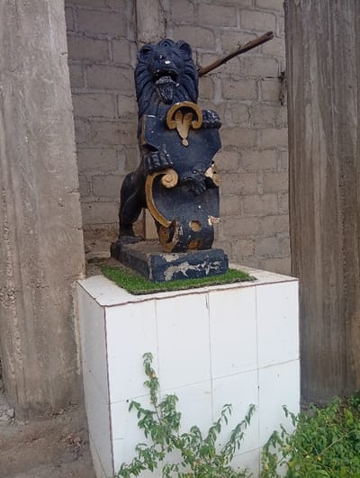 An outdoor scene in Itakpe Junction, Nigeria, depicts a weathered lion statue on a tiled pedestal. The black lion statue stands on its hind legs, featuring a detailed mane, open mouth, and bared teeth. It holds a dark-colored shield with gold-painted scrollwork, showing visible chips and deterioration. The statue rests on a small patch of artificial green turf atop a white-tiled pedestal with brown grout lines. Behind the statue is a light-colored concrete block wall. To the left and right, rough, unpainted concrete pillars flank the scene. A light brown wooden stick protrudes from the brick wall, angled upwards behind the lion's head. Green leafy vegetation grows from the base of the pedestal and on the unpaved ground in the foreground. No individuals are present.