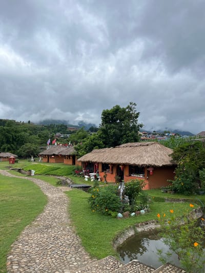 The image depicts a village with traditional houses, possibly in Ban Mai Saha Sampan, Thailand, given the context provided. The houses have orange walls and thatched roofs. A stone pathway winds through a grassy area in the foreground. In the background, a misty, mountainous landscape is visible, with some buildings scattered on the hillsides. Several flags are displayed on the rooftops of the houses. There are also decorative elements like lanterns and small ponds with water and flowers. The sky is overcast and cloudy, contributing to a subdued atmosphere. No people are visible in the scene.