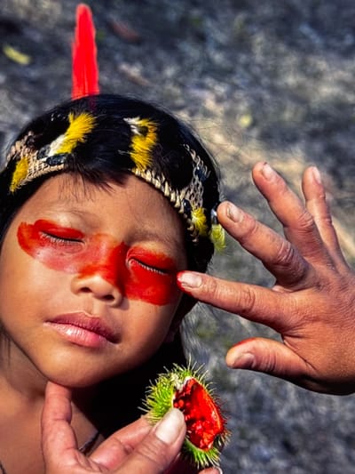 The image is a close-up portrait of a young child, possibly indigenous, adorned with red and yellow face paint across their eyes and a beaded headband with feathers. A hand, presumably an adult's, is positioned near the child's face, with fingers extended. The adult's hand is also holding a small, spiky green and red object. The background is blurred and appears to be natural terrain, with muted colors and textures suggesting an outdoor environment. There is no indication of the city of Orihuela, Spain, in the image.