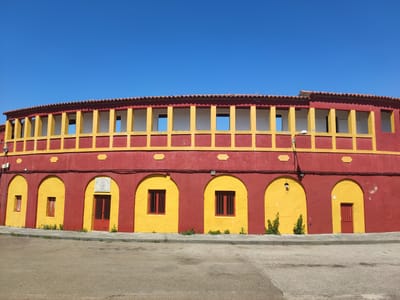 The image shows a circular building painted in red and yellow under a clear blue sky. The building has two levels. The lower level features a series of arched openings, some of which contain windows with red bars and some are red doors. The upper level has a long row of rectangular openings framed by yellow pillars and a red border. A tiled roof tops the structure. The building is situated on a paved street with a sidewalk in front of it, and some small green plants grow along the base of the building. A lamppost is visible on the right side of the building, attached to the wall. The building appears to be a bullring, located in Valencia de Alcántara, Spain.