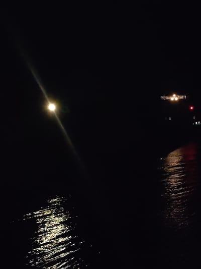 The image depicts a nighttime scene in what appears to be a waterfront area of Zvoz, Russia. The foreground is dominated by a dark body of water, presumably a river or lake, with visible light reflections suggesting ripples or waves. A bright light source, possibly a street lamp, illuminates the water's surface, creating a distinct light path towards the lower left of the image. In the distance, across the water, is a structure with multiple lights, possibly a pier, a bridge, or a building on the shoreline. A red light is also visible on the structure. The overall environment is dark and has an atmosphere of quiet calm. No people or other objects are visible in the image.