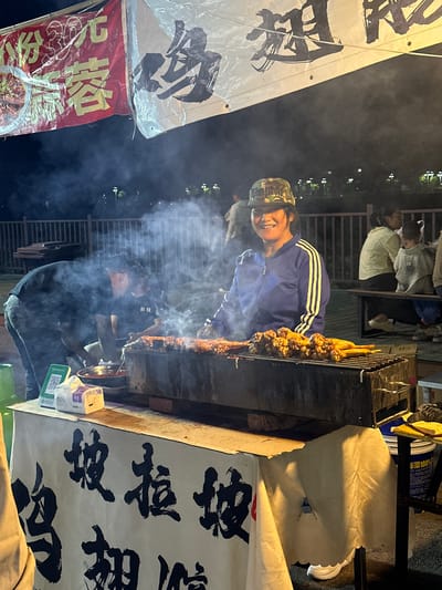 At a night market stall in Eshan Yi Autonomous County, China, a woman wearing a camouflage baseball cap and a blue and yellow striped jacket smiles at the camera while standing behind a grill. She is in front of a metal barbecue rack laden with skewered meats, which are emitting smoke. To her left, a person in a black t-shirt and jeans is tending to the grill. A white banner with black Chinese calligraphy is draped across the front of the stall. In the background, other people are seated on a bench, and a wooden fence runs along the perimeter of the market. The scene is illuminated by artificial light, creating a lively night market atmosphere.