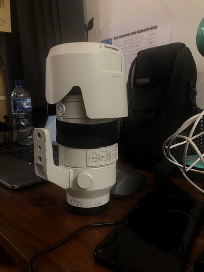 The image displays an indoor scene, likely a workspace or desk, with a large white camera lens as the central subject. The lens, equipped with a matching lens hood and a tripod collar, is positioned vertically on a wooden surface.
Visible text on the lens hood reads "ALC-SH145." On the lens barrel, controls for focus range "FULL 1 0.3m," optical steady shot ("ON II OFF"), and mode selection ("MODE 1 11 2") are discernible. The lens mount is identified as "E-mount," accompanied by regulatory symbols. The tripod collar attached to the lens shows signs of wear, with visible scratches.
Surrounding the lens, several other objects are present. A black camera bag is situated to the left of the lens, resting on a sheet of paper that appears to be a calendar or schedule with grid lines and partially legible text. White cables are tangled above and behind the lens and camera bag. To the right of the lens, a black tablet or smartphone screen lies next to a gray computer mouse. A laptop keyboard and a partially visible screen are beneath the lens, with a plastic water bottle, labeled "AQUA," placed on the keyboard. A book with green text is visible on the far left. The scene suggests a photographer's equipment and a cluttered yet functional workspace in Cibinong, Indonesia.