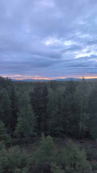 The image depicts a scenic landscape, likely captured during the golden hour. The left side of the frame is dominated by a vast expanse of clouds, painted with hues of lavender and pink, suggesting a sunset or sunrise. The clouds are textured and dynamic, with some appearing bright and illuminated, hinting at the sun's rays breaking through.

As the eye moves to the right, the sky transitions to a thin band of warm orange and yellow light along the horizon. This light illuminates a distant mountain range, creating a silhouette against the colorful sky.

The right side of the image is filled with a dense forest of dark green trees. The trees appear to be deciduous, with a variety of shapes and sizes, creating a textured wall of foliage. The overall impression is one of natural beauty and tranquility. The time of day is clearly indicated by the dramatic sky colors. The location, Khurmuli, Russia, suggests a wilderness setting. No people or objects are visible, and there is no visible text.
