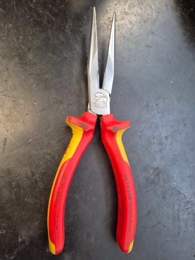 An overhead shot shows a pair of red and yellow insulated long-nose pliers lying on a dark, textured surface. The pliers are positioned vertically, with the tips pointing upwards. The metal jaws are visible at the top, slightly separated. The handles are predominantly red, with yellow accents on the grips. Text is visible on both handles: one handle reads "1000 V Made in GERMANY 14", and the other has "26 16 200 Vanadin Super". The background surface appears to be a workbench or countertop with some smudges and scratches, consistent with a workshop environment. There is no indication of a specific location within the city of Trier, Germany.