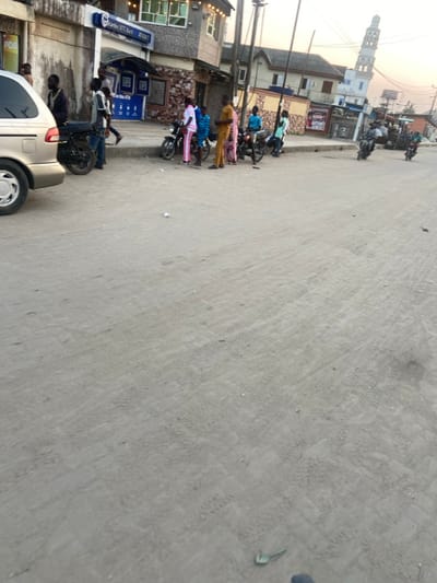A street view features a parked car and a row of motorcycles along a building with storefronts, including one that appears to be a bank. People are standing near the motorcycles. A tall mosque minaret rises in the background amidst other buildings. The road surface is grey and textured. The scene is captured in daylight, with a slightly hazy atmosphere.