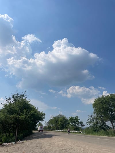The image shows a view in Katauhar Kalan, India, with a prominent sky filled with cumulus clouds. A paved road runs along the right side of the frame, bordered by trees. A vehicle is seen in the distance on the road. Debris and trash are visible along the roadside. A cyclist is visible on the road.