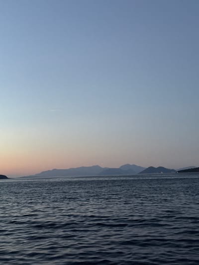 A horizontal, medium shot captures the sea and distant mountains against a gradient sky at dusk. The water in the foreground is dark and choppy with visible ripples, reflecting the dim light. A range of subdued, blue-hued mountains forms the horizon line, with a faint pink and orange glow on the sky just above them, transitioning to a pale blue higher up. The scene is peaceful and serene. The location is the sea near Vathy, Greece. There are no people, animals, or man-made structures clearly visible.
