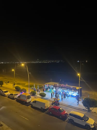 An aerial shot shows a bustling street scene at night. A snack shop is lit brightly, drawing a crowd of people. Cars of various colors are parked along the road adjacent to the shop. The image's background is dark, illuminated by streetlights stretching into the distance, suggesting a town or city along the coast. Vegetation and green areas are visible along the road.