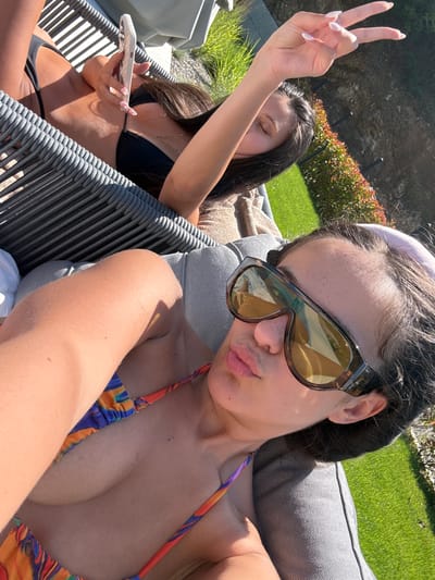 This outdoor photograph depicts two young women relaxing in Elenovo, Bulgaria, under bright daylight. The woman in the foreground, positioned closer to the camera, has dark hair and wears large, dark brown sunglasses with amber lenses. She is sporting a colorful bikini top with a vibrant pattern in hues of blue, orange, and purple, and exhibits a slightly puckered lip expression, suggesting a selfie pose. She reclines on a light gray cushioned outdoor lounge chair.
Behind her, partially visible, is another woman with dark hair. Her eyes appear closed or downcast, and she is wearing a black bikini top and bottom. She holds a white smartphone in one hand and raises her other hand, forming a peace sign, revealing manicured nails with light polish. This woman is seated on a gray woven outdoor chair.
The background shows a sunny outdoor environment with bright green grass, well-kept bushes featuring reddish-brown foliage, and a natural, rocky hillside further back. The strong sunlight casts clear shadows, indicating a bright, clear day, likely midday or early afternoon. The overall scene conveys a relaxed, leisurely atmosphere, characteristic of a vacation or poolside setting. No text is visible in the image.