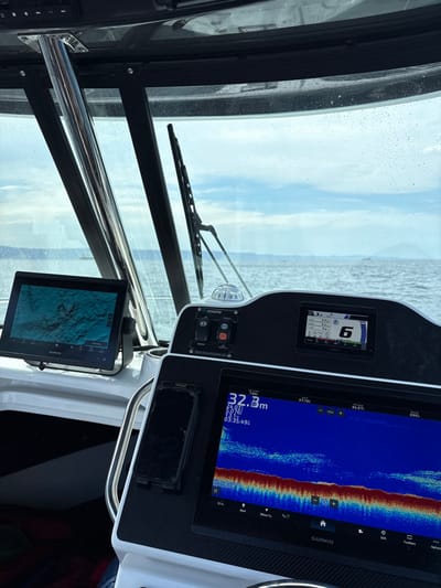 The boat's dashboard features a windshield with a wiper, a digital display showing speed, a depth finder with readings at 32.3 meters, and a tablet displaying a nautical chart. Water droplets obscure the windshield, hinting at a wet environment. The dashboard has a carbon fiber texture and various controls. The background shows a blurred ocean view.