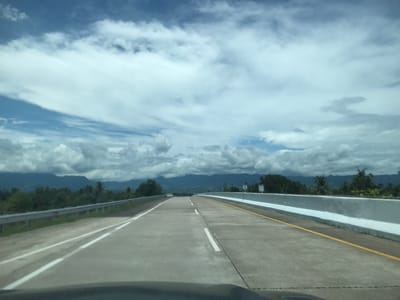 The image depicts a view from a vehicle driving on a highway in Cotkareung, Indonesia. The road is a concrete multi-lane highway with a center divider and yellow lines marking the right side. A gray metal guardrail is on the left side, and a concrete barrier is on the right. A green sign with "KM 55" is visible on the right side of the road. In the background, there are green trees and hills under a partly cloudy sky. The sky is primarily blue with white clouds. Part of the dashboard of the vehicle is visible at the bottom of the image.