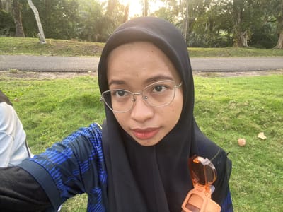 The image is a selfie of a young woman wearing a black hijab and glasses. She is outdoors, likely in a park or natural setting with green grass and trees visible. A portion of a black and blue striped shirt is visible on her right arm. In the top left corner, an orange object, possibly sunglasses or a small container, is attached to her hijab. The lighting suggests it is daytime, with sunlight filtering through the trees, creating a warm glow. The background shows a paved path, more grass, and dense foliage with palm trees. The woman's expression is neutral, with her eyes looking directly at the camera. The orientation of the image is rotated 90 degrees clockwise.