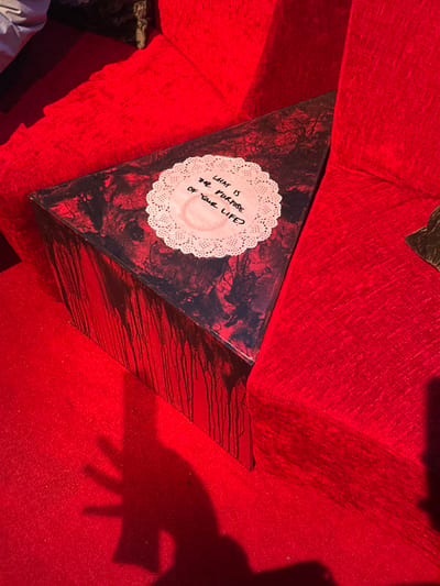 The image displays an interior scene, presumed to be in the City of Westminster, United Kingdom, characterized by a dominant red color scheme. A dark, roughly triangular prism-shaped object with irregular, textured surfaces is centrally positioned. Vertical dark, glossy streaks, resembling dried paint drips, run down the sides of this object. A white, circular paper doily with a scalloped edge rests on the object's top surface. Handwritten in black ink on the doily are the words "WHAT IS THE PURPOSE OF YOUR LIFE?", beneath which a faint, light brown circular stain, consistent with a cup mark, is visible. The object is situated on a bright red, textured carpet or rug, and is flanked by sections of red upholstered furniture, likely a sofa or chair armrests. A distinct shadow of a human hand, with fingers extended, is cast onto the red carpet in the lower left portion of the frame. No individuals are directly visible.