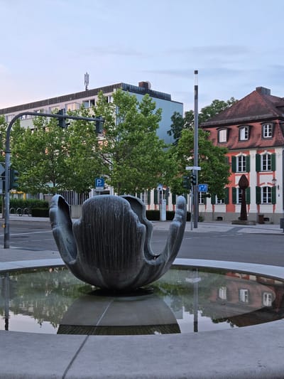 This is a ground-level, medium-shot, eye-level photograph of a modern sculpture in a fountain, situated in the city of Ansbach, Germany. The sculpture, made of dark, textured metal, resembles a large, open seed pod or a stylized flower. It is partially submerged in the still water of a circular fountain, with its upper portions rising above the water's surface. The water reflects the surrounding environment, including the sky, trees, and buildings.
In the background, on the left side of the image, is a multi-story modern building with a flat roof. A cell tower is mounted on top of this building. In front of this building, trees with lush green foliage are visible. Traffic lights and street signs are also present.
To the right of the modern building, a traditional, older building with a red tiled roof and white and red painted facade is visible. This building has multiple windows with green shutters. In front of this building, a tall, slender, dark-colored statue or monument stands.
The scene is set at dusk, with a soft, diffused light illuminating the sky, which transitions from pale blue at the top to a lighter hue near the horizon. The overall impression is one of an urban public space, likely a plaza or square, in Ansbach.