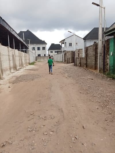 The scene shows a dirt road with a single person walking away from the camera. The person is wearing a green shirt and blue jeans. The road is flanked by concrete walls and houses. The houses are two-story buildings with white walls and brown roofs. There are streetlights on the side of the road, and the sky is overcast.  The location is Ovwian, Nigeria. The coordinates (latitude: 5.485, longitude: 5.806) place the location in the city of Ovwian, Nigeria. The scene is a typical example of an unpaved road in a residential neighborhood in Nigeria. There are no other people in the scene. The road is dusty and the houses look newly built.