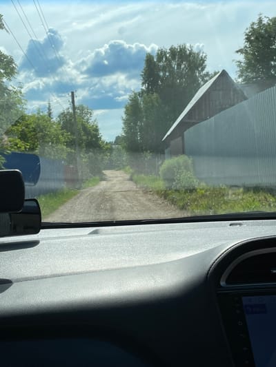 The image captures a view from inside a vehicle, looking out through the front passenger window on a bright, sunny day. The camera is oriented vertically, showing the scene as if it were viewed with the head tilted.

Outside the window, a bright blue sky with fluffy white cumulus clouds dominates the upper half of the frame. Green trees with dense foliage are scattered across the landscape, with some appearing closer and others further in the background. A utility pole with power lines is visible against the sky and trees on the left side. Running parallel to the vehicle is a dirt or gravel path, flanked by green grass and more trees. A building with a dark, corrugated metal roof is partially visible in the distance, appearing behind the trees.

Inside the vehicle, the textured gray plastic of the car's dashboard and door panel is visible on the right side, mostly in shadow, indicating strong sunlight from the left. An infotainment screen or a similar digital display is partially visible in the upper right corner, showing some interface elements and the number "440". Several black objects, possibly accessories or parts of the car's interior, are seen in the lower right portion of the image. Reflections of the car's interior are faintly visible on the window glass, blending with the exterior view.

The scene depicts a daytime setting, likely mid-day given the brightness and cloud formation, in a rural or suburban area identified as Пятый километр, Russia.