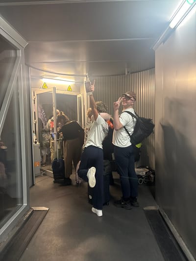 Two young men pose in a corridor. The first man wears a hat, a backpack, and gives a peace sign. The second man points. Both wear white shirts and dark pants. Other people stand behind a glass partition to the left, where warning symbols are visible. Luggage is scattered on the corridor floor. The space appears to be an airport or train station.