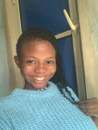This image, captured in Onibudo, Nigeria, displays a frontal portrait of a female subject, estimated to be a young adult. She possesses dark skin and black box braids, with her hair styled away from her forehead. The subject is smiling broadly, revealing her teeth, and maintaining direct eye contact with the camera. She is dressed in a light blue, chunky knit sweater. A garment with a visible black and white animal print pattern is worn underneath, its collar peeking out at her neck. Her left hand is visible on the right side of the frame, lightly grasping a strand of her braided hair. The background indicates an indoor setting, with a textured, light-colored wall occupying the left portion of the frame. To the right, a darker blue surface, consistent with a door or window pane, is visible, featuring a white, curved handle/latch assembly.