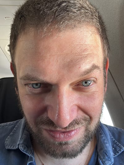 A close-up portrait shot depicts an adult Caucasian male, positioned from the forehead to below the chin, looking directly at the camera. He possesses short, dark-blonde to light-brown hair with visible grey strands, particularly along the hairline. His blue eyes are wide open, and his mouth exhibits a subtle upward curve, forming a smirk. The subject has a short, trimmed beard and mustache. His skin appears fair, with a reddish tone on the cheeks and nose. Horizontal lines are present on his forehead. He is attired in a blue denim-style collared shirt, beneath which a thin gold-colored chain necklace is visible. The background is blurred, indicating an indoor environment, with elements in the upper left suggesting an overhead compartment or paneling typical of an aircraft cabin. This scene is located in Peschiera Borromeo, Italy.