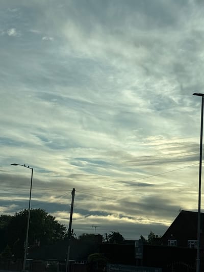 The image shows a street scene in South Staffordshire, United Kingdom, under a cloudy sky. The sky is filled with a variety of clouds, ranging from wispy cirrus to more dense stratus formations. There are two streetlights visible, one on the left and one on the right side of the frame. The streetlight on the left has three birds perched on its arm. Below the sky, there are houses and trees, with some power lines crisscrossing the scene. The overall impression is that of an overcast day, possibly late afternoon or early morning, given the quality of light.