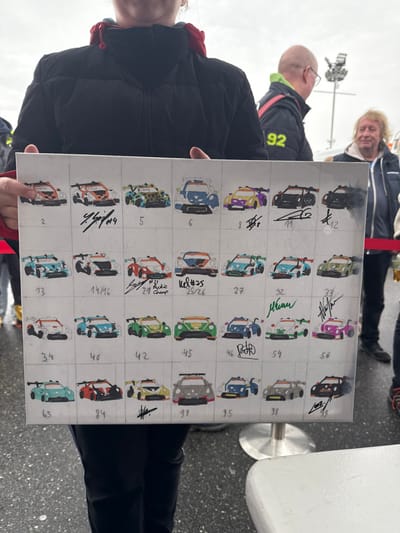 An outdoor scene at a motorsport venue in Hockenheim, Germany, under an overcast sky. A person, largely obscured from the neck down and wearing a dark puffer jacket with a red collar, holds a rectangular white canvas board horizontally. The board displays a grid of approximately 42 cells (6 rows x 7 columns visible) each containing a stylized, brightly colored illustration of a race car. Many cells are numbered (e.g., 2, 4, 5, 6, 8, 11, 12, 13, 14/16, 25, 27, 32, 33, 34, 40, 42, 45, 46, 54, 56, 65, 84, 91, 95, 98, 99). Numerous cells feature black ink signatures and additional text, such as "Y. Schi #4," "J.Sch #21 Rookie 24 Champ," and "ud #25 25/26." In the background, to the right, a bald man with glasses, wearing a dark jacket with a yellow-green collar and "92" on the back, looks left. Further right, an older man with light curly hair, wearing a dark blue jacket, stands looking forward. A red barrier tape runs horizontally across the mid-ground. The ground is dark asphalt or concrete. A partial white table with a silver pedestal base is visible in the lower right foreground. Metal support structures are discernible in the far background against the grey sky.