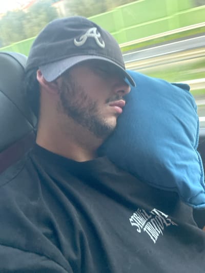 A young adult male is depicted in a close-up, side-profile view, appearing to be asleep or resting. His head is tilted to the right, supported by a light blue pillow. He wears a dark-colored baseball cap with a light grey brim and a prominent white "A" logo, consistent with the Atlanta Braves sports team. Dark hair is visible beneath the cap, and he has a beard or heavy stubble. His lips are slightly parted. He is wearing a black t-shirt featuring a partially visible white graphic design on the left chest, which includes stylized text. The immediate environment includes dark, possibly leather, seat upholstery behind his head and shoulder. Through a window in the background, a heavily motion-blurred outdoor scene is visible, characterized by indistinct green foliage and light-colored horizontal streaks, indicating movement within a vehicle. There is no visual information present in the image to determine a specific location within the city of Leiria, Portugal.