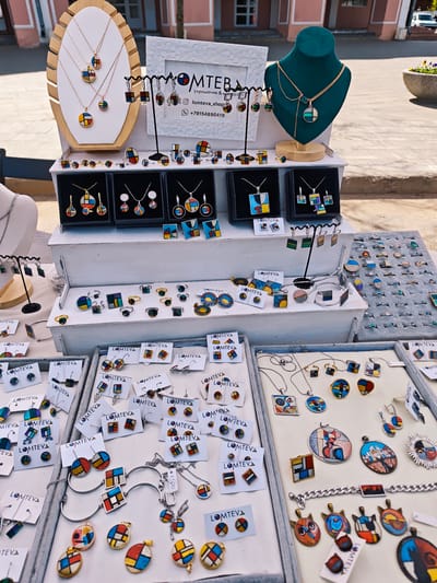 The image shows a jewelry display at an outdoor market or event in Uglich, Russia. The jewelry, which appears to be handmade, features a modern, geometric design with colorful enamel or resin inlays. A sign displays the name "LOMTEVA" along with contact information, suggesting it is the brand or seller's name. Jewelry is arranged on multiple tiers and trays, including necklaces, earrings, rings, and bracelets. A green velvet bust displays a necklace, while a wooden display showcases other necklaces. The jewelry has a distinct style, and it is artfully displayed to attract customers. The setting is likely a street fair or craft market within the city.