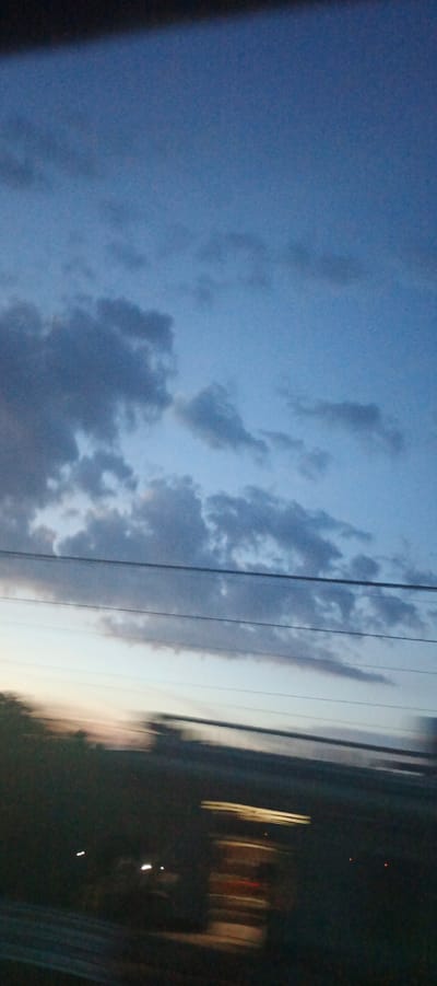 The image captures a dusk or dawn sky with scattered clouds, transitioning from a deep blue overhead to a lighter hue near the horizon. Three parallel wires stretch horizontally across the frame, obscuring a sliver of the sky. Below the wires, the landscape is blurred and dark, suggesting motion, possibly from a vehicle. Indistinct lights, appearing as streaks, are visible in the lower right portion of the image, indicating structures or vehicles in the distance. The overall scene appears to be an exterior view, likely taken from a moving vantage point within or near the city of Xuân Long, Vietnam, given the context provided. There are no discernible people, specific subjects, or explicit actions.