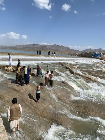 The image shows a group of people at a dam or weir, with mountains in the background under a bright blue sky with scattered clouds. The location is identified as Kim, Afghanistan. Several men are standing in and around the water flowing over the structure. Some are fully clothed, while others are shirtless or partially dressed. One man is looking at a phone. The water is rushing over the stepped stone surface of the dam, creating white water. A sign is visible on the right side of the structure, with text in a non-English script.