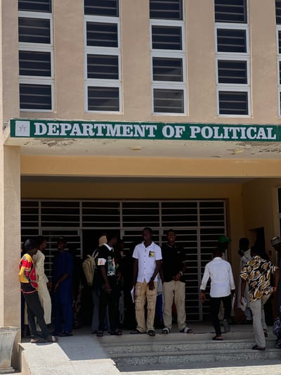 The photograph captures a group of young men standing outside a building, under a sign that reads "DEPARTMENT OF POLITICAL". The building appears to be a public institution, possibly a university or college, given the presence of students. The architecture is simple, with beige walls and rows of windows covered by horizontal slats. The group is gathered on a set of steps leading up to an entrance. The lighting suggests it is daytime, likely outdoors under a bright sun. The men are dressed in casual attire, with some wearing traditional African clothing and others in Western-style shirts and pants. Their expressions and stances indicate they are engaged in conversation or waiting for something. The overall impression is one of everyday life at an educational institution. While the city of Yali, Nigeria is mentioned, the image itself does not contain any explicit visual cues to confirm this specific location.