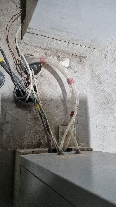 The image displays an electrical wiring installation on a dusty concrete floor in Chapyk, Uzbekistan. No people are visible in the frame.
The central focus is a cluster of electrical conduits and wires connecting two gray, metallic electrical cabinets or panels. Numerous clear or translucent flexible plastic conduits, bundled with red tape or ties at various points, emerge from circular openings in the concrete floor. They arc upwards and lead towards penetrations at the base of the right-hand cabinet. Visible inside these conduits are various electrical wires, identifiable by colors such as red, white, and green. A specific green wire with a distinct yellow piece of insulation or a connector is also prominent among the cables.
On the left side, exposed red and white wires, along with darker, possibly black, conduits or wire bundles, emerge from another opening in the concrete floor, heading towards the left electrical cabinet. The wires generally appear to be routing power or data between the floor and the electrical enclosures.
The scene suggests an ongoing construction or renovation activity, or a utility area. The concrete floor is rough and heavily coated in dust and debris, indicating an active work environment or an area not yet finished. The lighting indicates an indoor setting. There is no visible text within the image. The overall impression is that of a complex electrical connection point under development.