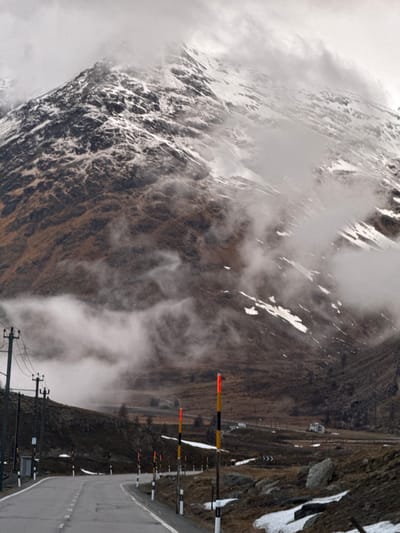 The image depicts a rugged, high-altitude mountain pass, likely in the Swiss Alps near Pontresina, under overcast and misty conditions. When viewed correctly (rotated 90 degrees clockwise from the provided image), a paved road with a white dashed center line stretches from the foreground into the distance on the lower right side, gently curving. It is lined on its right by a series of tall, distinctive road markers, some with alternating orange and black segments, others white and black, likely snow poles or avalanche indicators. Further to the right of the road, utility poles with power lines run parallel to the asphalt.
To the left of the road, a massive, steep mountain face dominates the scene, heavily textured with rocky outcrops and significant patches of snow. Swirling clouds and thick mist partially obscure its peaks and higher slopes, creating a dramatic and atmospheric vista. On the right side of the road, another mountain slope rises, covered in a mix of sparse, brownish vegetation, rocks, and intermittent snow patches.
The weather is cold and damp, characterized by the persistent low clouds and visible snow. The diffuse light indicates an overcast day, contributing to the cool and subdued palette of greys, browns, and whites. No people or visible text are present, emphasizing the wild and remote nature of the alpine environment. The scene evokes a sense of solitude and natural grandeur.